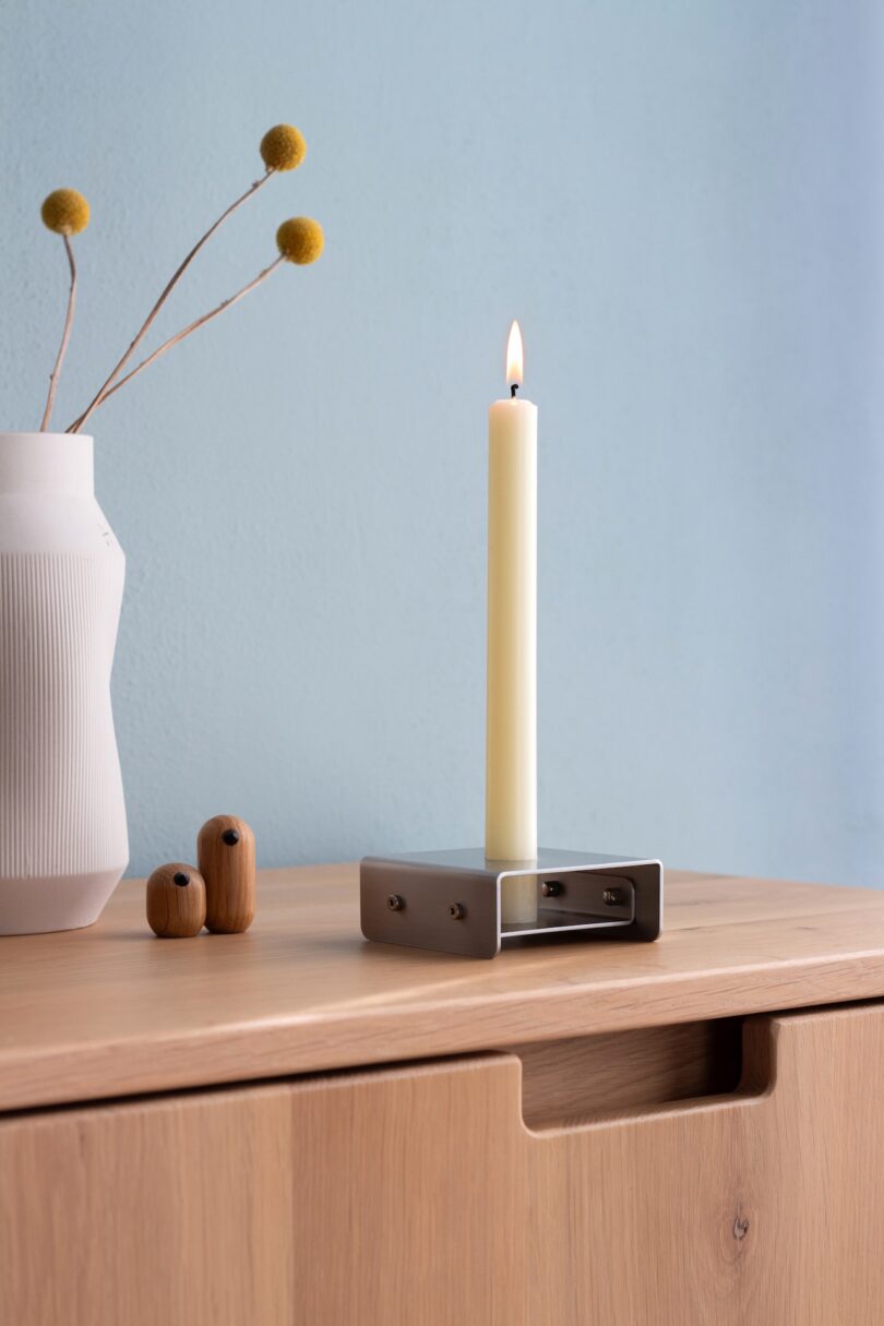 A lit candle in a metal holder sits on a wooden surface next to a white vase with yellow billy balls and two small wooden decorative objects.