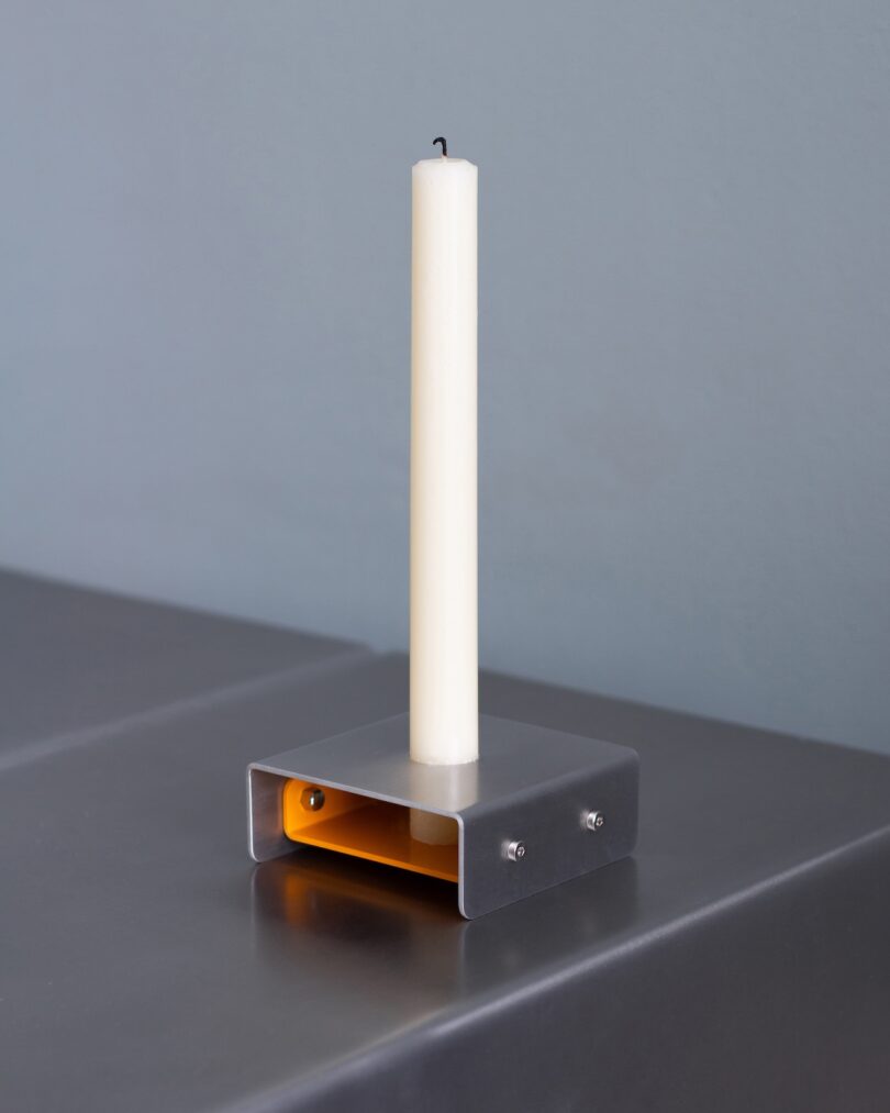 A white taper candle is placed in a minimalist square metal holder with an exposed yellow interior, set on a gray surface against a gray wall.