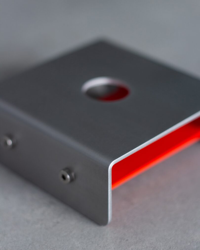 A close-up of a metal bracket with a red interior, featuring a central circular hole and screws on the side, placed on a gray surface.