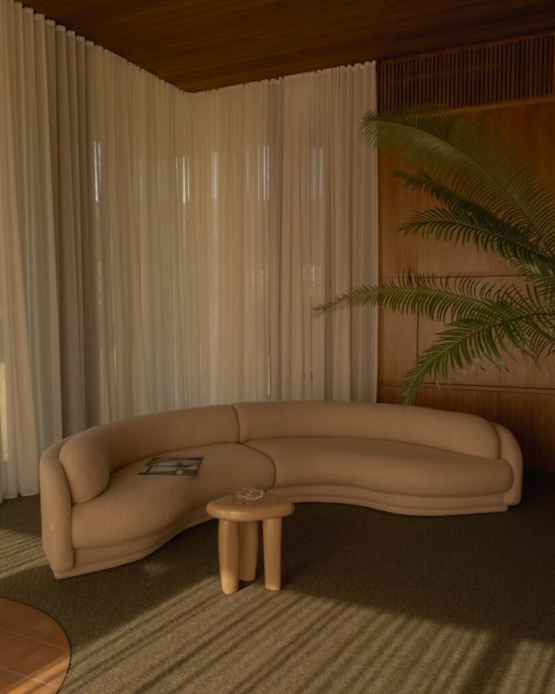 A beige curved sofa with a magazine, a small round wooden table, and a potted plant in a room with sheer curtains, wood panel walls, and carpet flooring