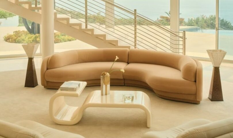 A modern living room with a curved beige sofa, a sculptural white coffee table, two geometric lamps, and large windows overlooking an outdoor pool and staircase