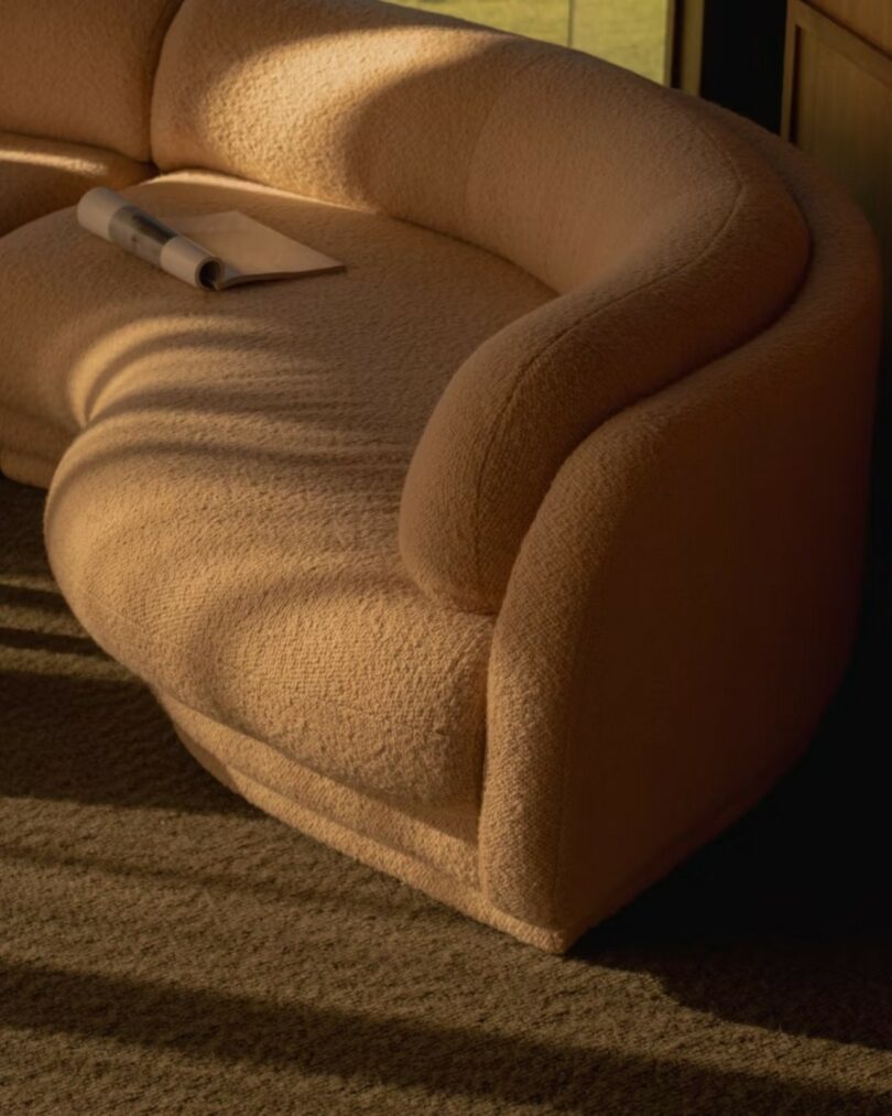 A beige curved sofa sits on a carpeted floor in sunlight, with a rolled-up newspaper and paper resting on one seat cushion