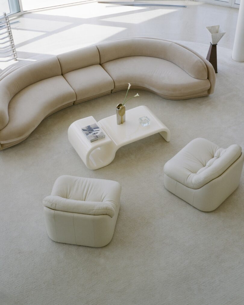 Curved beige sofa, two matching armchairs, and a white coffee table with a vase, magazine, and glass item arranged in a minimalist, light-filled living room