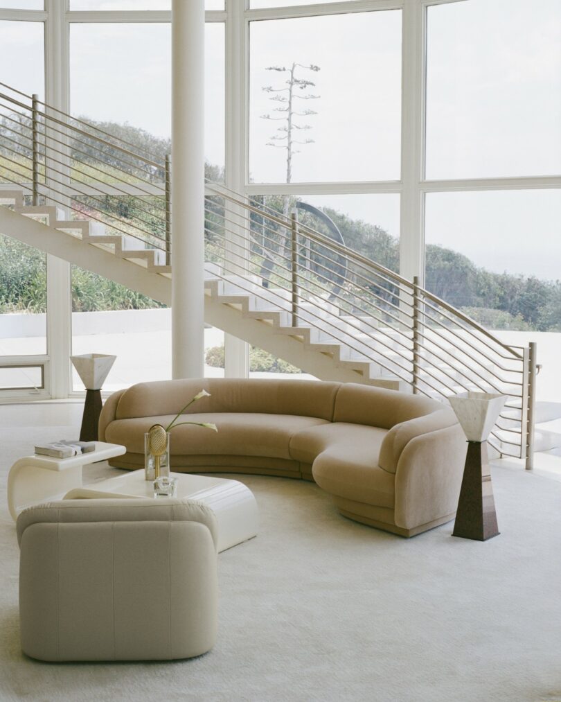 Modern living room with a curved beige sofa, white armchair, two marble lamps, a small table, and large floor-to-ceiling windows near a metal staircase