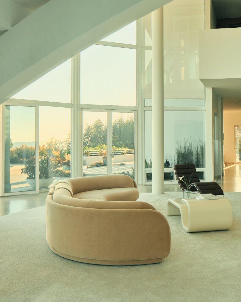 Modern living room with large windows, a beige curved sofa, two black chairs, and a white coffee table overlooking an outdoor garden