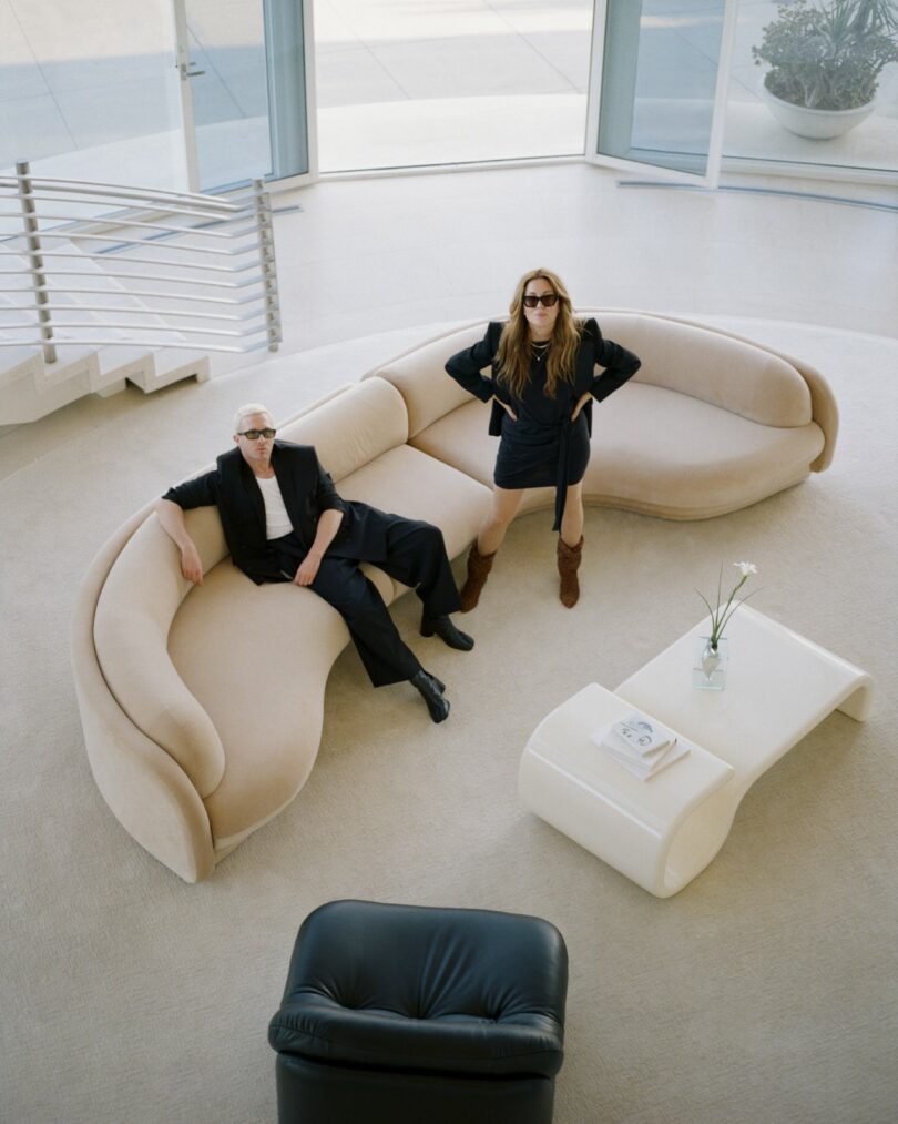 Two people wearing dark clothing and sunglasses are posed on and beside a curved beige sofa in a modern, bright living room with large windows and minimal decor