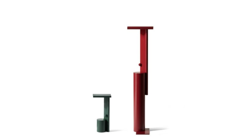 Two minimalist, abstract sculptures in red and green stand side by side against a plain white background. The taller sculpture is red, and the shorter one is green
