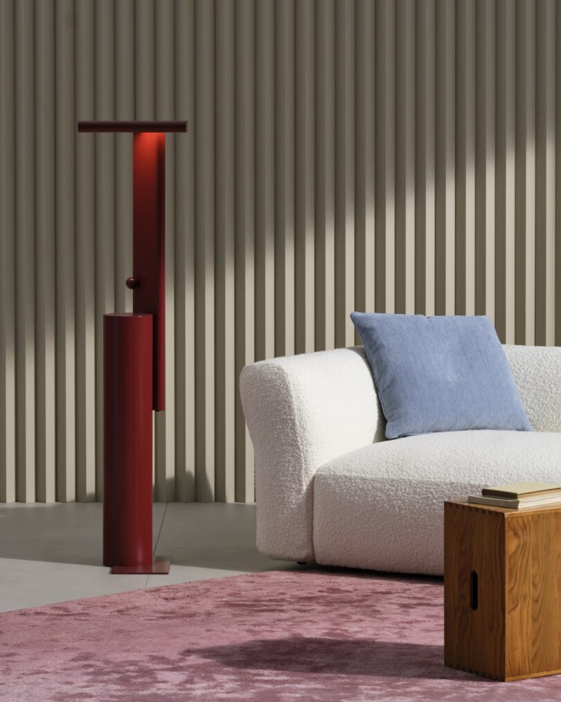 A modern living room with a white textured sofa, blue pillow, wooden side table, red floor lamp, pink rug, and beige vertical wall panels