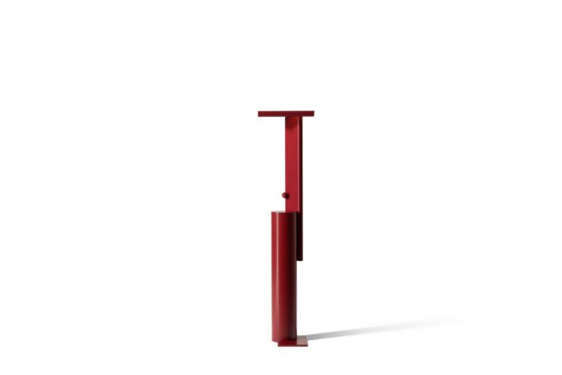 A minimalist red metal object with a vertical cylindrical base and a flat horizontal top, standing against a plain white background