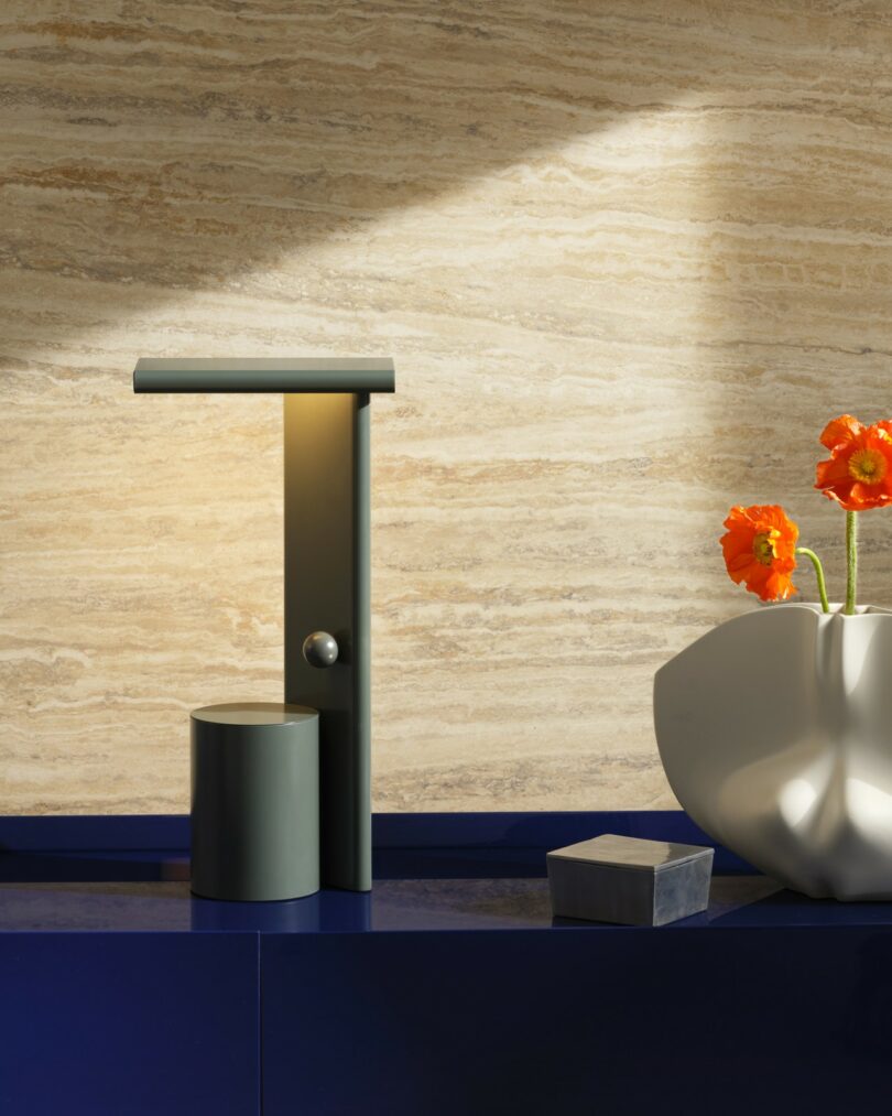 A modern lamp, a sculptural white vase with orange poppies, and a small gray box are arranged on a glossy blue surface against a beige stone wall