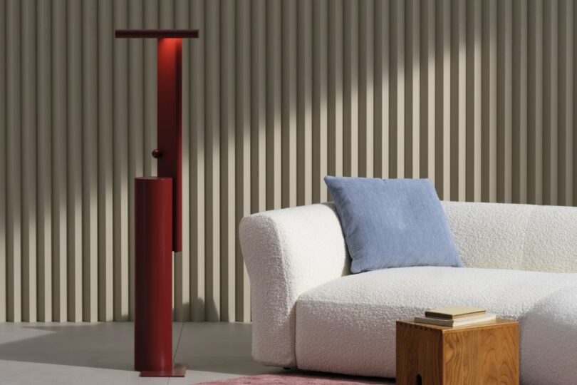 A modern living room features a white textured sofa with a blue pillow, a small wooden table with books, a red floor lamp, and a ribbed beige wall in the background