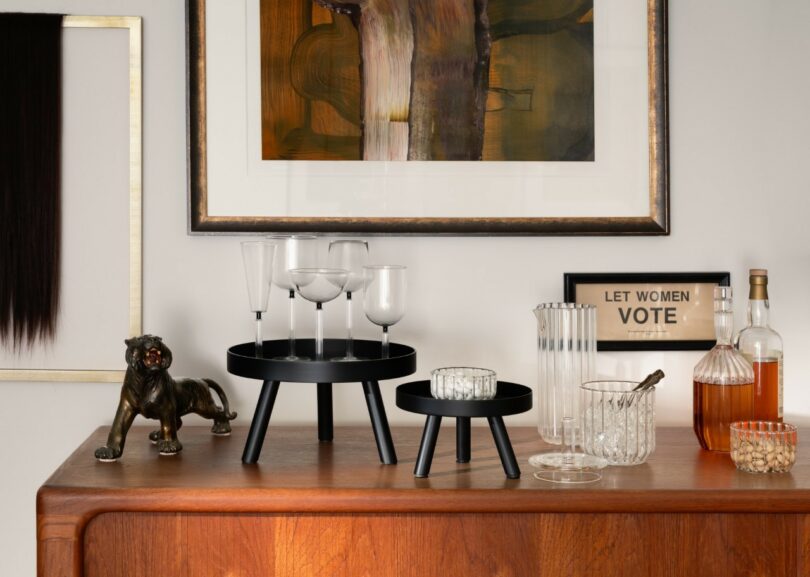A wooden sideboard with glassware, a decanter, bottles, a black panther figurine, and a framed “LET WOMEN VOTE” sign beneath abstract artwork