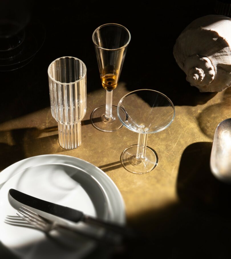 Three empty glass cups, a white plate with a fork and knife, and a large seashell are arranged on a brass surface under warm light
