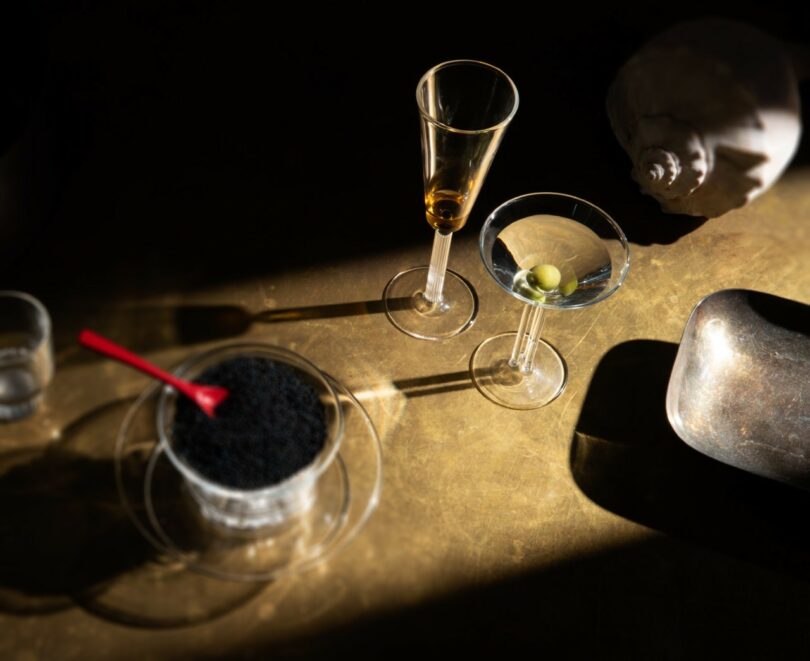 A glass of brown liquor, a martini with an olive, a bowl of black caviar with a red spoon, a metal scoop, and a seashell arranged on a sunlit surface