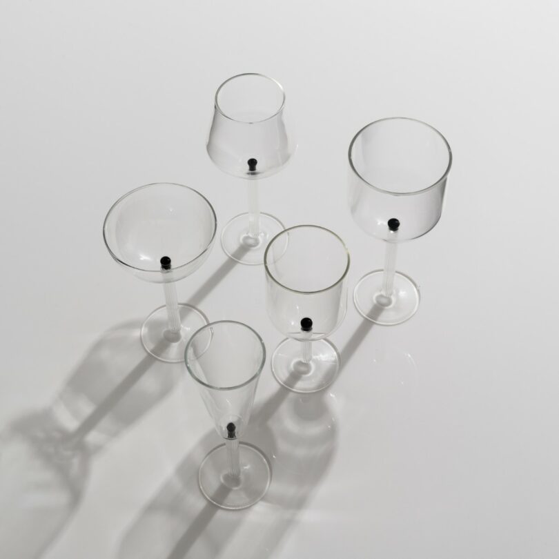 Five empty clear wine glasses viewed from above, each with a single small black dot at the base, casting soft shadows on a white surface