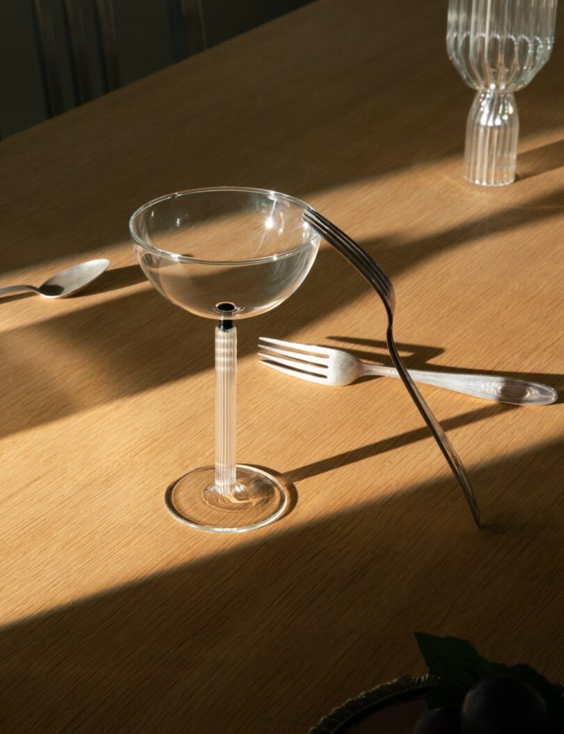 A clear cocktail glass and a fork balanced on its rim sit on a wooden table, with another fork, spoon, and glass nearby in sunlight
