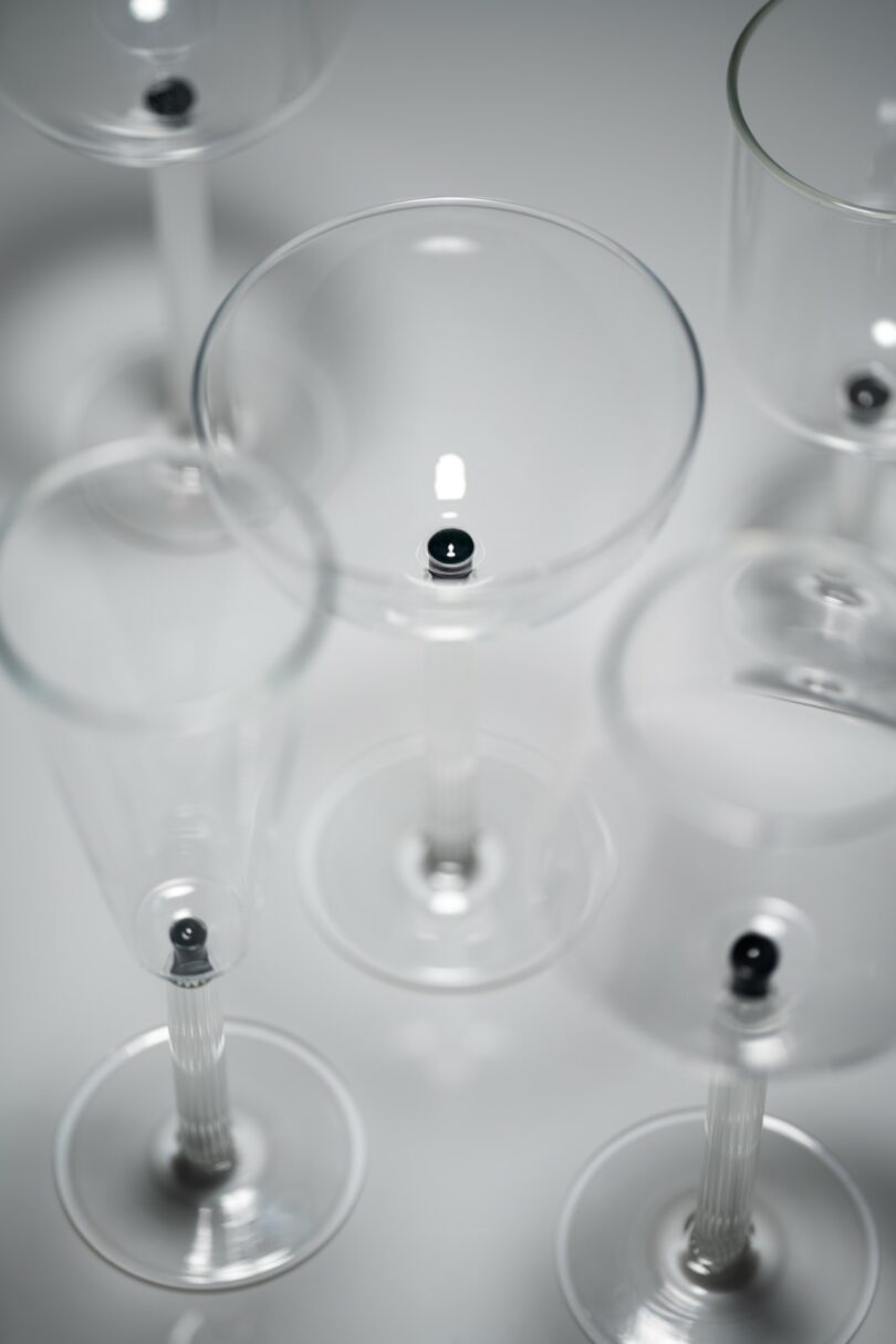 Several empty clear wine glasses are arranged upright on a white surface, each containing a single small black ball at the center of its base