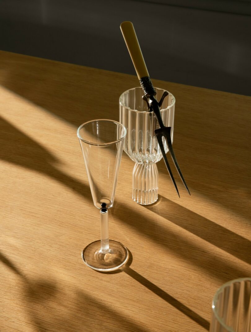 A clear wine glass, a ribbed glass cup, and a carving fork with a light handle are arranged on a wooden surface with shadows cast by sunlight