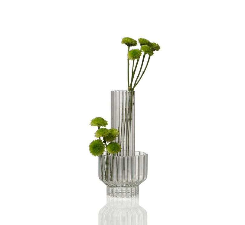 A clear, ribbed glass vase with two connected sections holds green pom-pom flowers, set against a white background