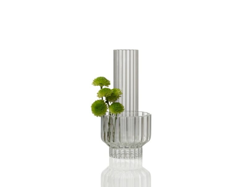 A clear, ribbed glass vase with vertical lines holds a few green pompom flowers, set against a plain white background