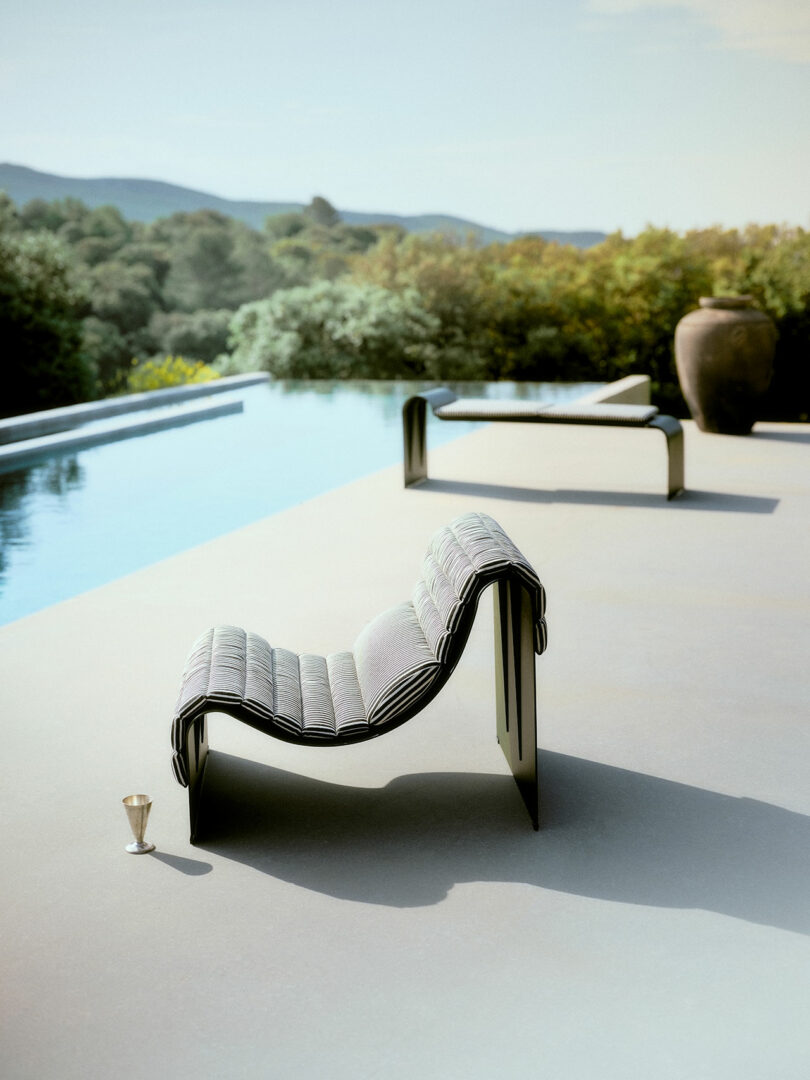 Two modern lounge chairs sit beside an infinity pool with a drink placed nearby; trees and hills are visible in the background under a clear sky