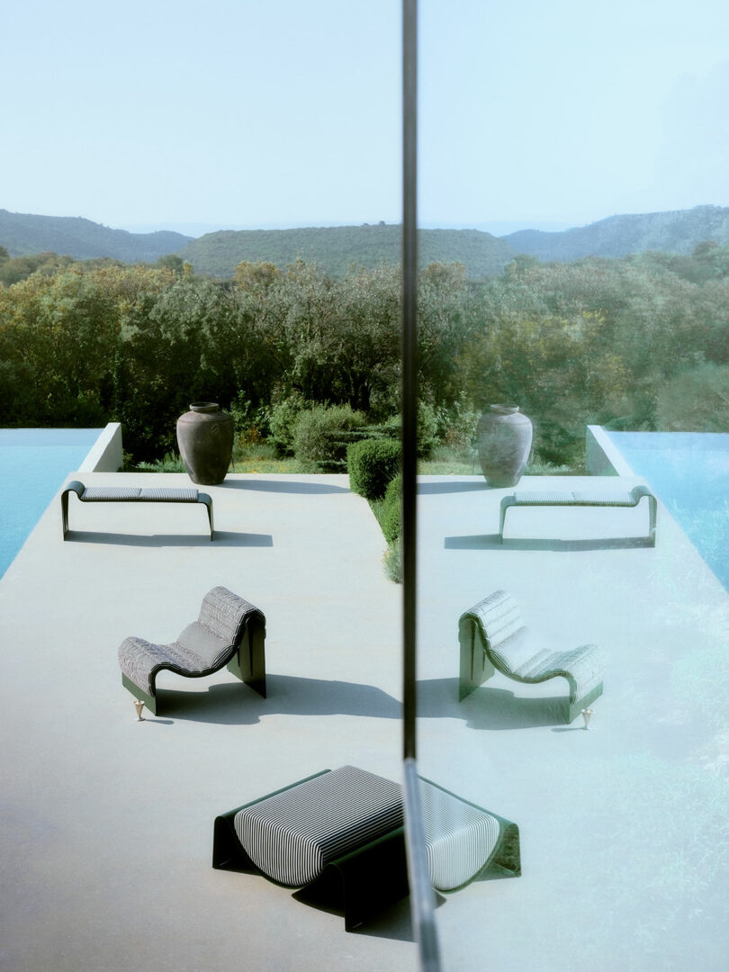 Modern poolside patio with lounge chairs and two large vases, reflected symmetrically in a glass wall, set against a natural landscape with distant hills and trees
