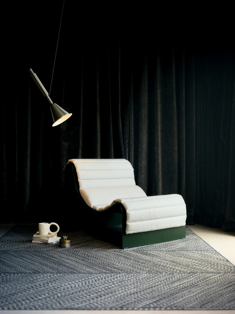 A modern lounge chair with a white cushioned surface on a dark rug, beside stacked books, a cup, and a saucer, under a hanging lamp, against dark curtains