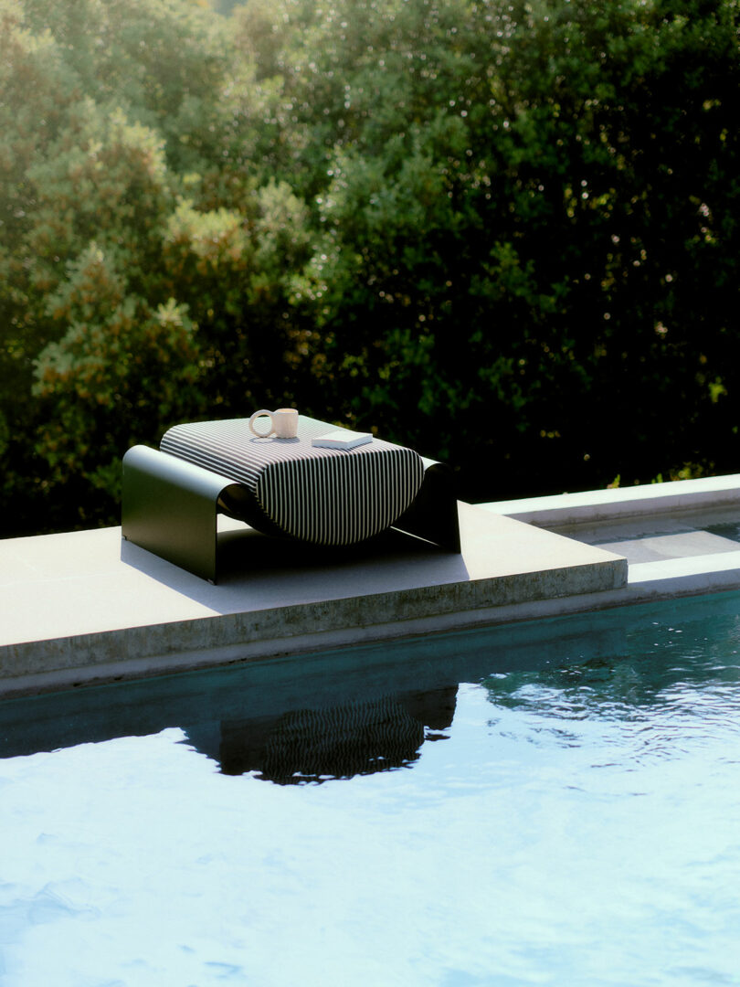 A modern black lounge chair with a striped cushion and a mug placed on top sits beside a pool, with green trees in the background