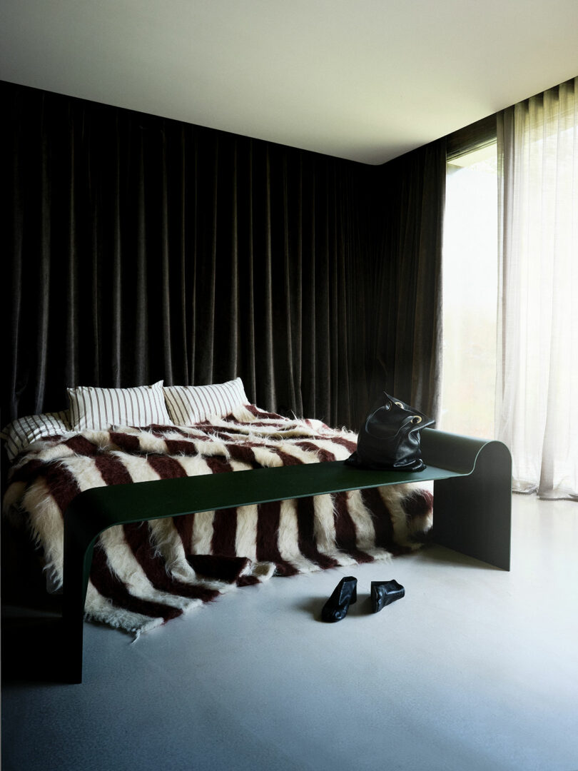 A bed with striped pillows and a fur-patterned blanket sits by a black bench with a black bag; black shoes are on the floor near sheer curtains and dark velvet drapes