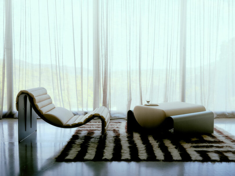 Minimalist living room with a modern lounge chair, a low table set on a patterned rug, and sheer curtains letting in natural light