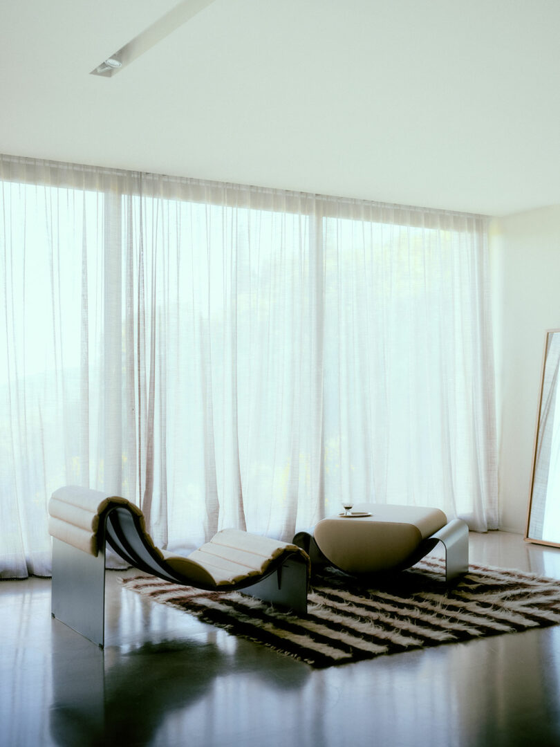 Modern minimalist lounge chair and matching ottoman on a zebra-patterned rug in front of sheer floor-to-ceiling curtains, with a drink and a mirror nearby