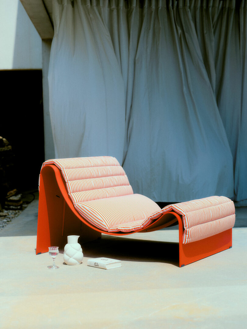 A modern red lounge chair with striped cushions and a matching footrest sits on a concrete patio, accompanied by a white vase, a book, and a glass; gray curtains hang in the background