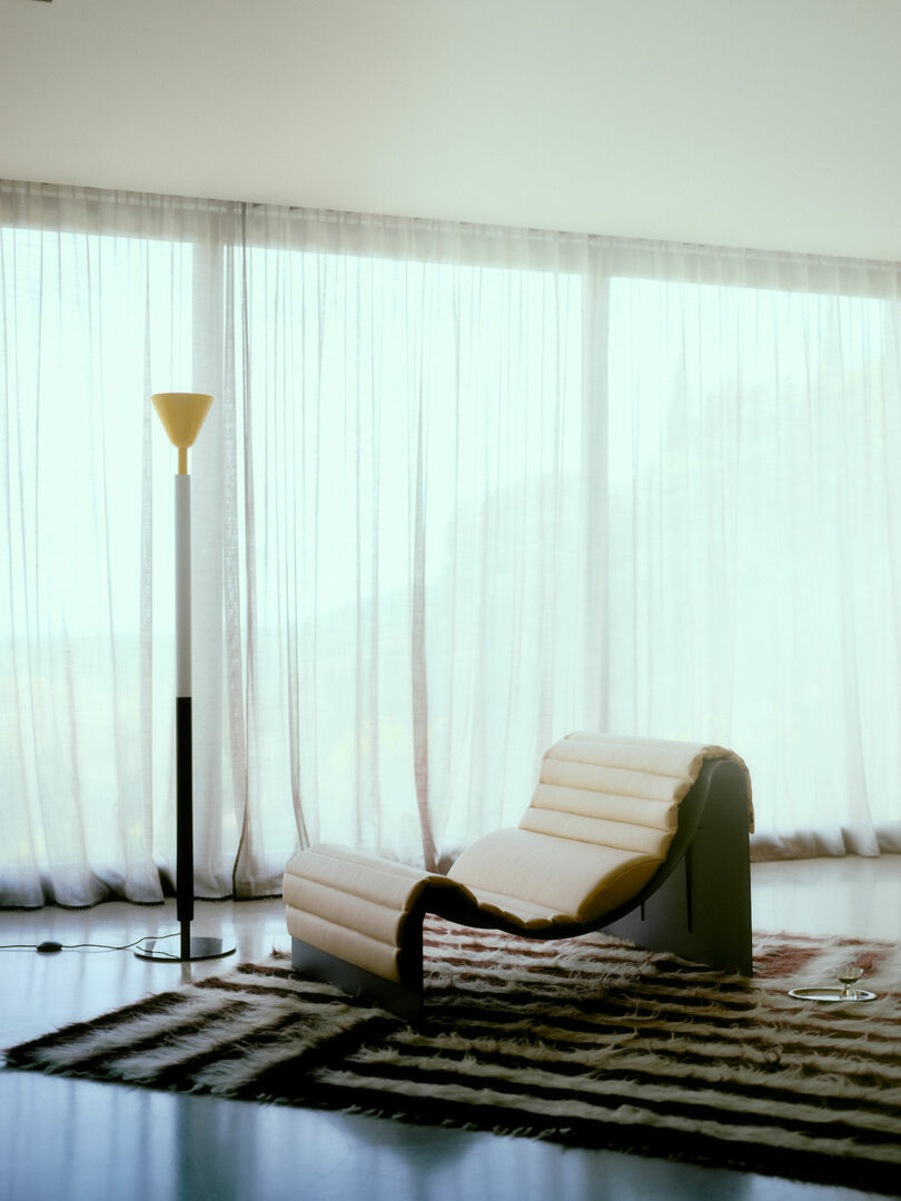 A modern lounge chair sits on a patterned rug next to a tall floor lamp in front of large, sheer-curtained windows
