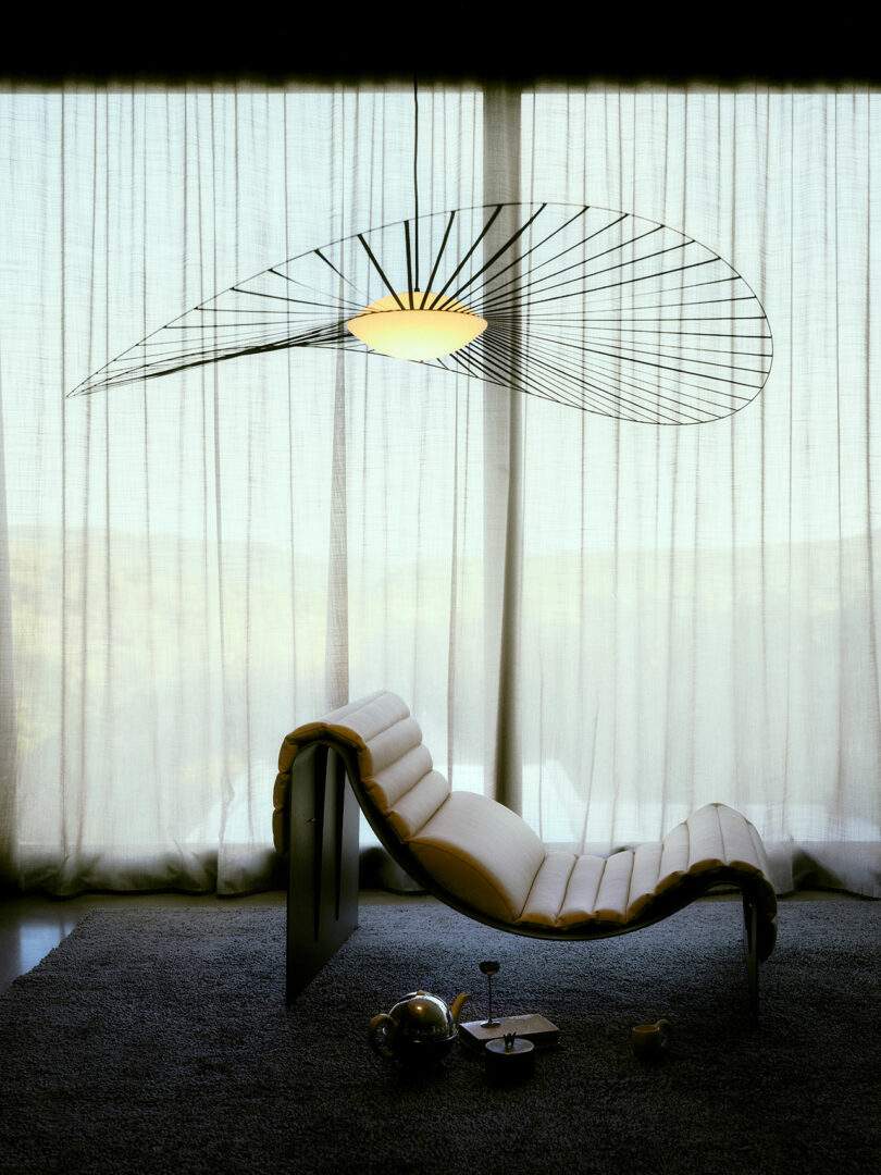 A modern lounge chair sits on a dark rug in front of sheer curtains, with a sculptural ceiling light above and a tea set placed on the floor nearby