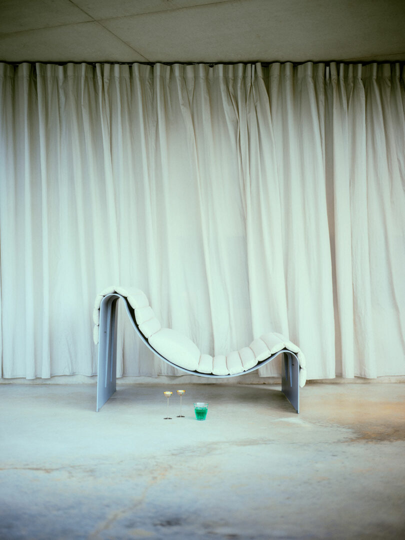 A modern white lounge chair sits on a concrete floor in front of white curtains, with two champagne glasses and a green drink placed in front of it