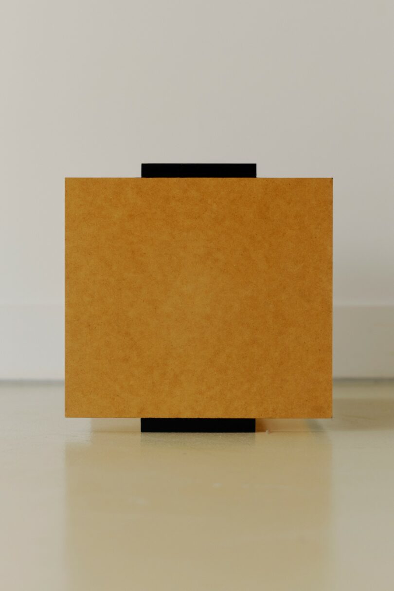 A rectangular brown board stands upright on a light floor, with a thin vertical black object partially visible behind it against a plain white wall