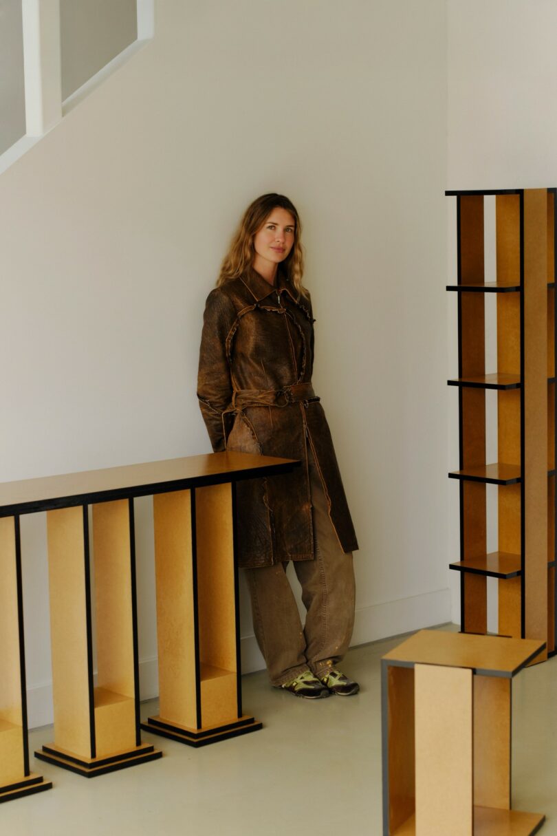 A woman in a brown coat and pants stands indoors beside modern, geometric wooden furniture pieces