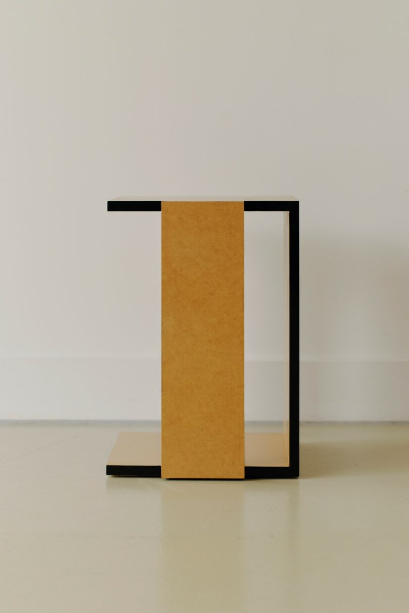 A minimalist, geometric side table with a black frame and a vertical rectangular tan panel, placed on a light-colored floor against a plain wall