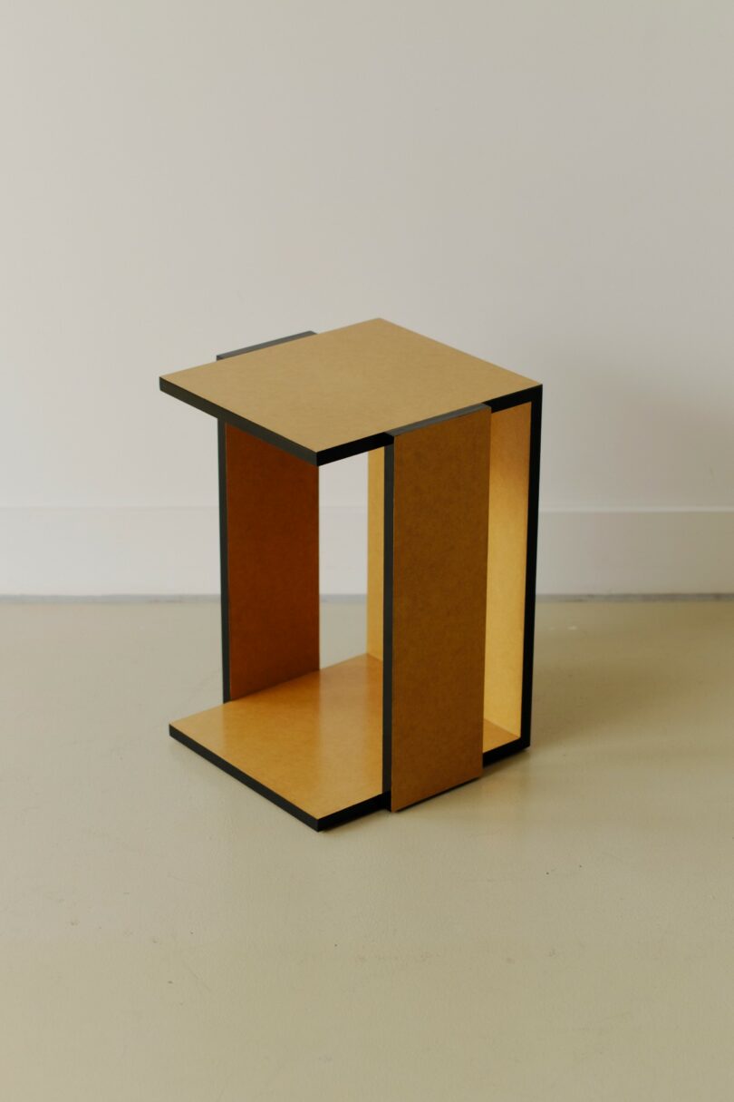 A small, minimalist wooden side table with an open rectangular design, placed on a light-colored floor against a plain white wall