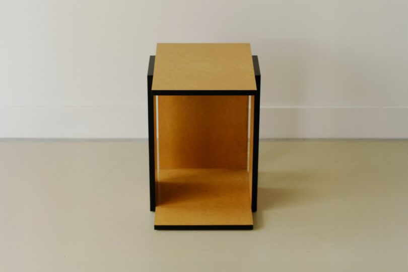 A small, rectangular, minimalist wooden table or stool with open sides, placed on a light-colored floor against a plain white wall