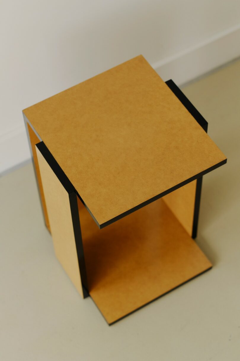 A modern, geometric side table with a light wood finish and black edges, positioned on a beige floor against a white wall