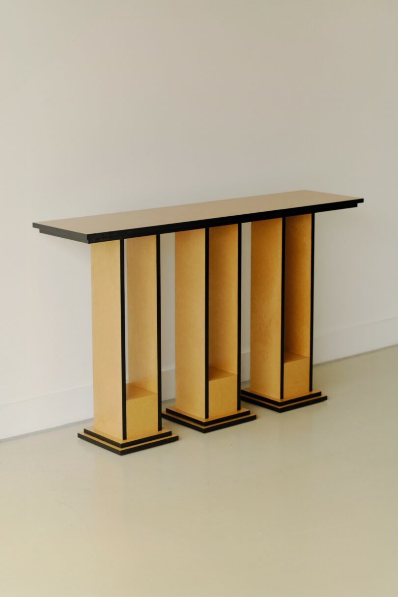 A modern console table with three rectangular columns, light wood panels, and black trim, set against a plain white wall and floor