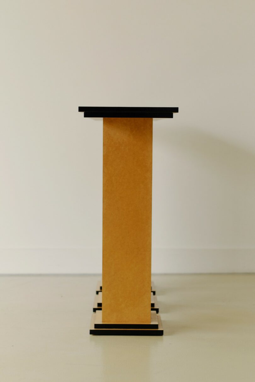 A rectangular, vertical structure with a black top and base, set against a plain white wall and floor, resembling a minimalist pedestal or table