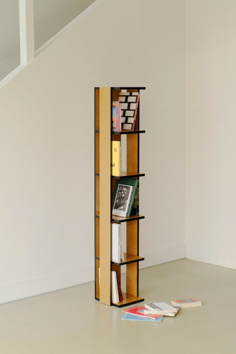A tall, narrow bookshelf with several books placed haphazardly on its shelves and a few books lying on the floor nearby, set against a plain white wall