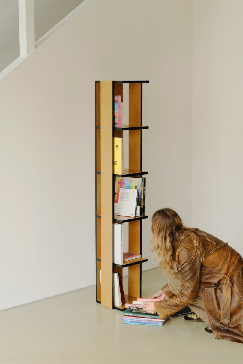 A person in a brown coat kneels on the floor, arranging books near a tall, narrow bookshelf against a light-colored wall
