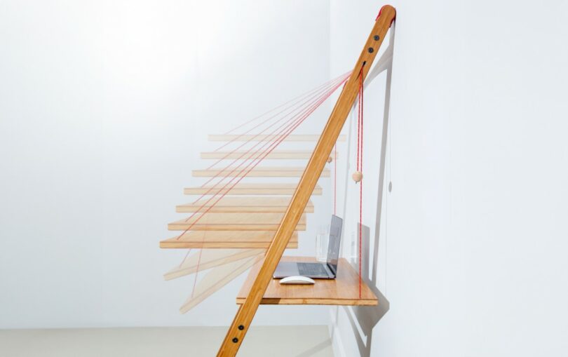 A wooden folding desk attached to a wall, with red strings and a laptop and mouse placed on the surface