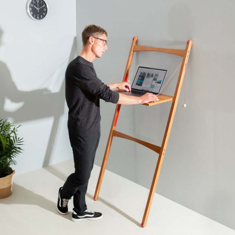 A man uses a laptop on a wooden standing desk that leans against a wall; a clock and a potted plant are visible in the room