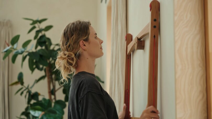 A woman with tied-back hair stands indoors, holding a wooden wall-mounted desk, with a large green plant in the background.
