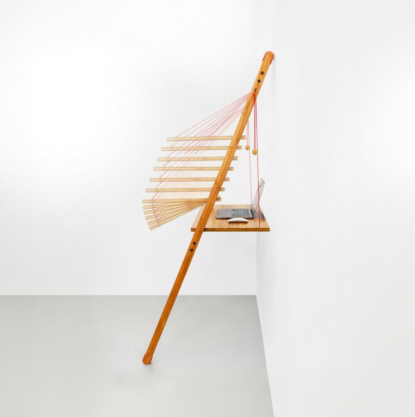 A wooden structure leans against a white wall, featuring horizontal wooden slats, red strings, and a small shelf holding a book and a dark bowl