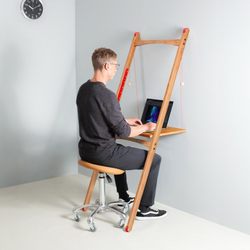 A man works on a laptop at a minimalist wooden desk leaning against a wall, seated on a wheeled stool. A clock is visible on the wall